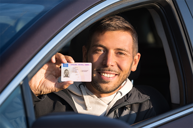 Smiling Latino man sitting in a car and holding his driver licence.